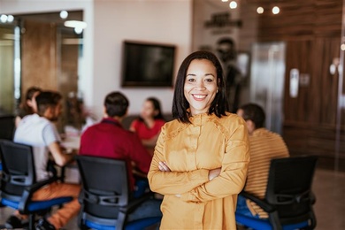 Modern office setting with a team collaborating around a conference table. A person stands confidently in the foreground wearing a mustard-yellow top, highlighting leadership and presence.