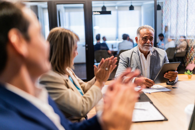 Business professionals seated around a conference table during a meeting, with one participant reviewing information on a tablet while others applaud, in a modern office setting with glass walls and colleagues working in the background.