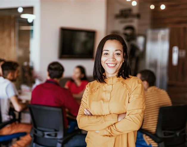 Modern office setting with a team collaborating around a conference table. A person stands confidently in the foreground wearing a mustard-yellow top, highlighting leadership and presence.