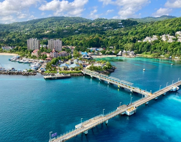 A vibrant coastal scene featuring a turquoise-blue bay with a long pier extending into the water. The shoreline is lined with resorts, marinas, and lush green hills in the background, creating a picturesque tropical setting under a partly cloudy sky.