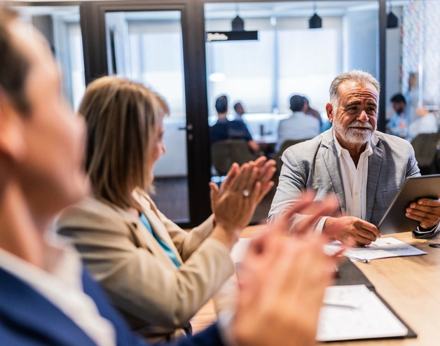 Business professionals seated around a conference table during a meeting, with one participant reviewing information on a tablet while others applaud, in a modern office setting with glass walls and colleagues working in the background.