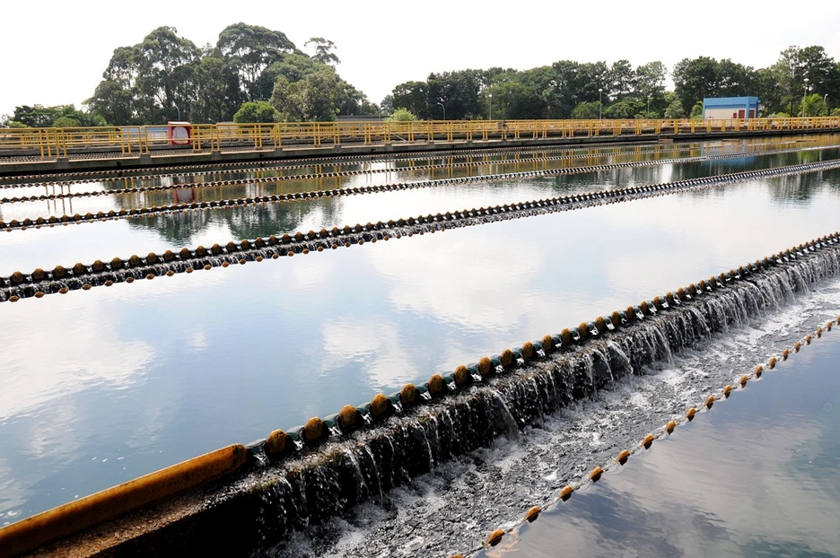 A wide view of a water treatment plant, with multiple parallel concrete channels where water flows evenly over stepped weirs. Yellow safety railings run along the walkways above the channels, and trees frame the background under a clear sky reflected in the water.